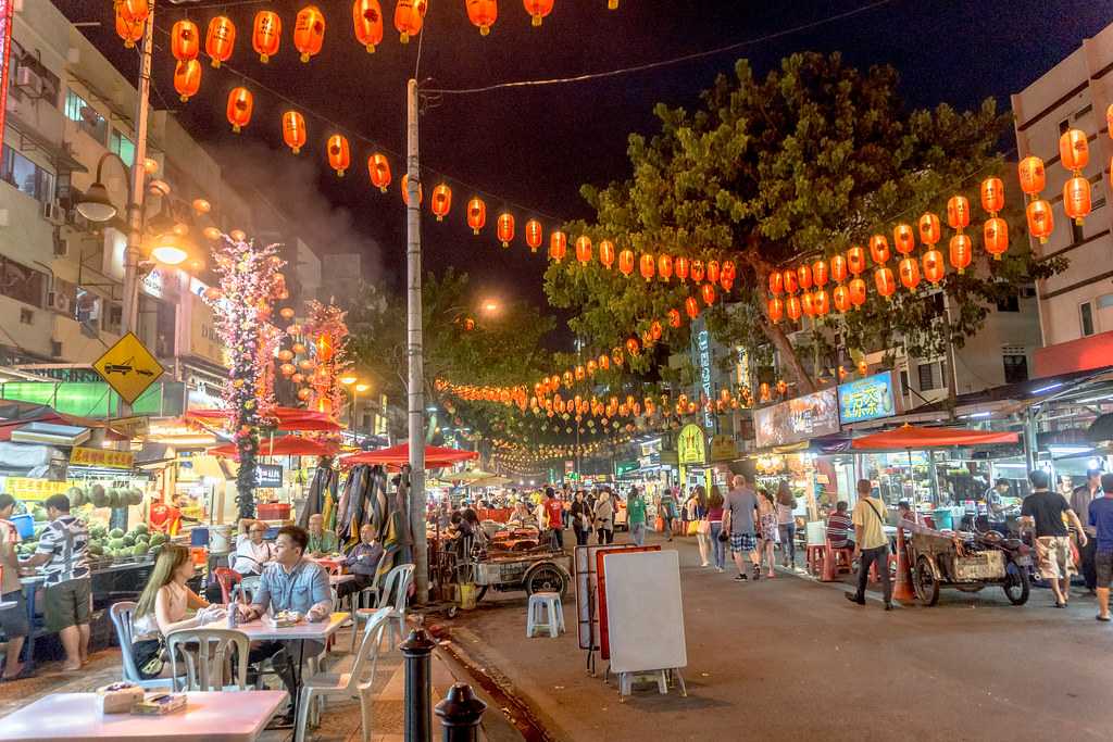 Puestos de comida callejera iluminados en Jalan Alor, Kuala Lumpur, durante la noche, con luces de colores y vapor.